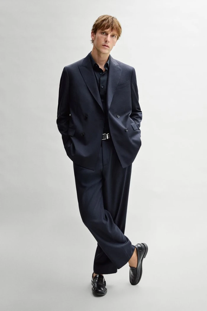 Model Quentin Demeester wearing an oversized navy double-breasted suit and black loafers against a light grey studio background.