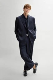 Model Quentin Demeester wearing an oversized navy double-breasted suit and black loafers against a light grey studio background.
