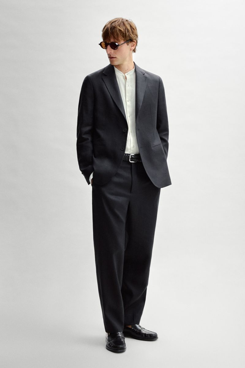 Quentin Demeester in a charcoal suit, cream band-collar shirt, and sunglasses against an off-white seamless background.