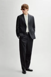 Quentin Demeester in a charcoal suit, cream band-collar shirt, and sunglasses against an off-white seamless background.