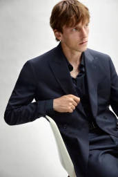 Model Quentin Demeester seated in a navy double-breasted suit against a clean white studio backdrop.