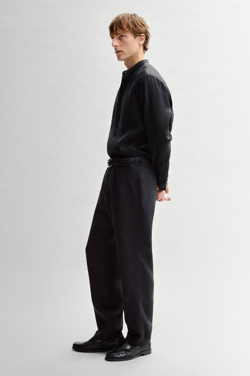 Model Quentin Demeester posing in profile wearing an all-black shirt and tailored trousers against an off-white seamless backdrop.