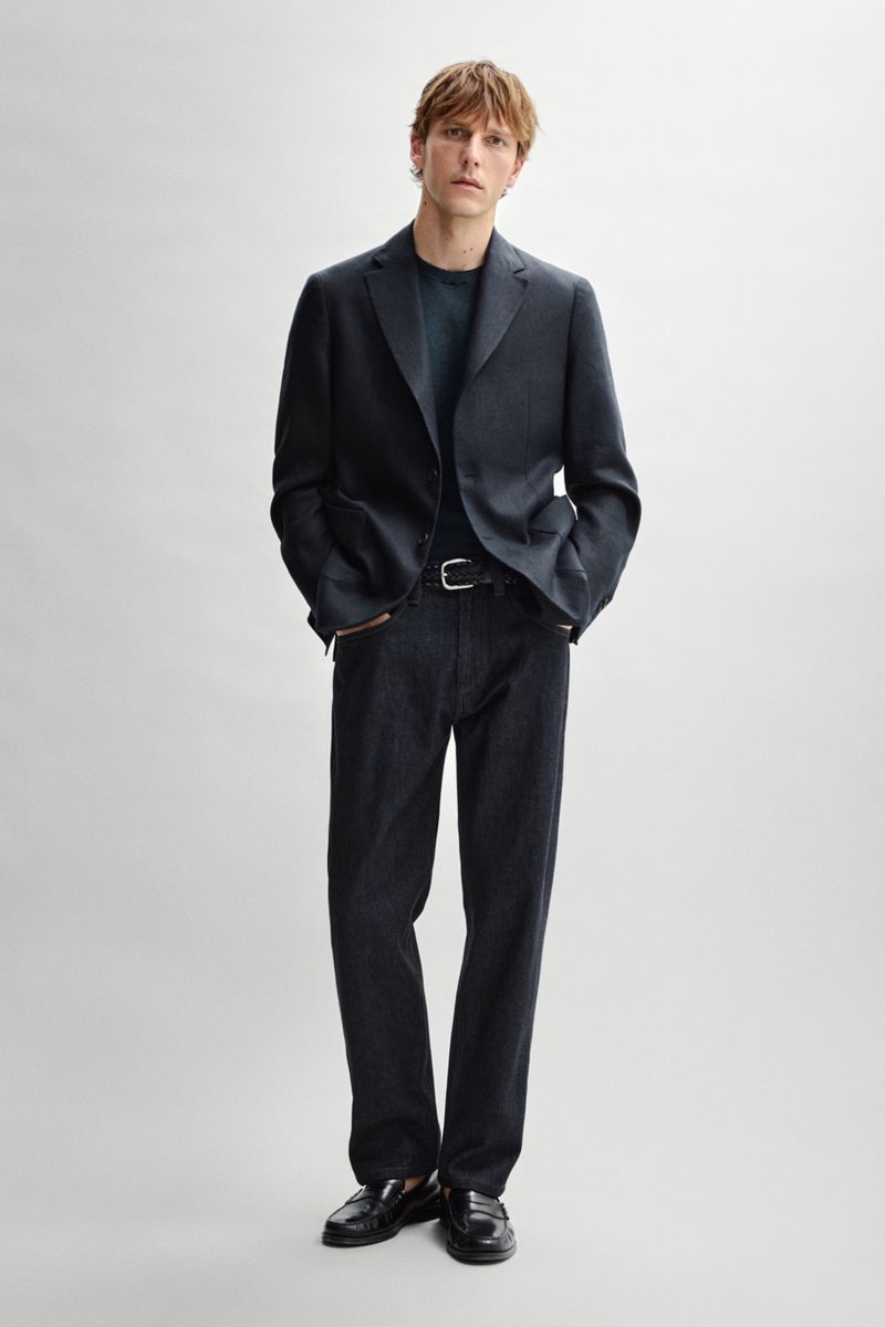Model Quentin Demeester standing in a dark tailored blazer, dark denim trousers, and black loafers against a light grey studio background.