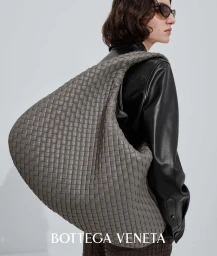Model wearing a black leather jacket carrying an oversized grey woven leather intrecciato shoulder bag against a plain studio background.