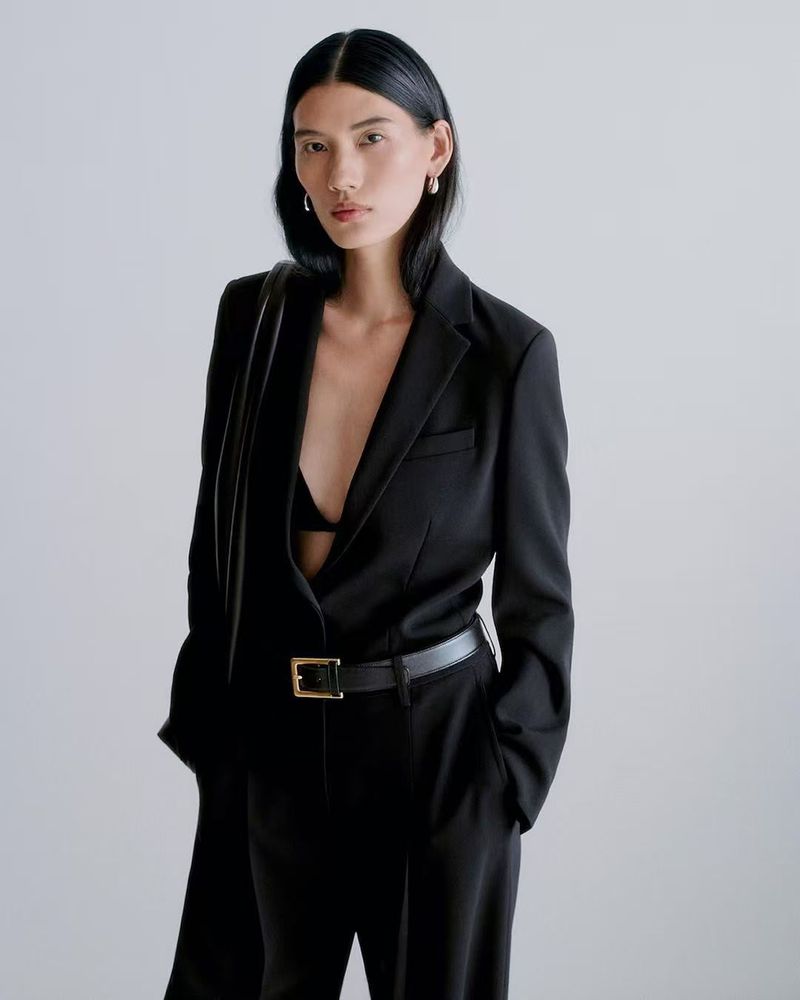 Model Jiayun Xie poses in a tailored black suit with a black leather belt against a neutral studio background.