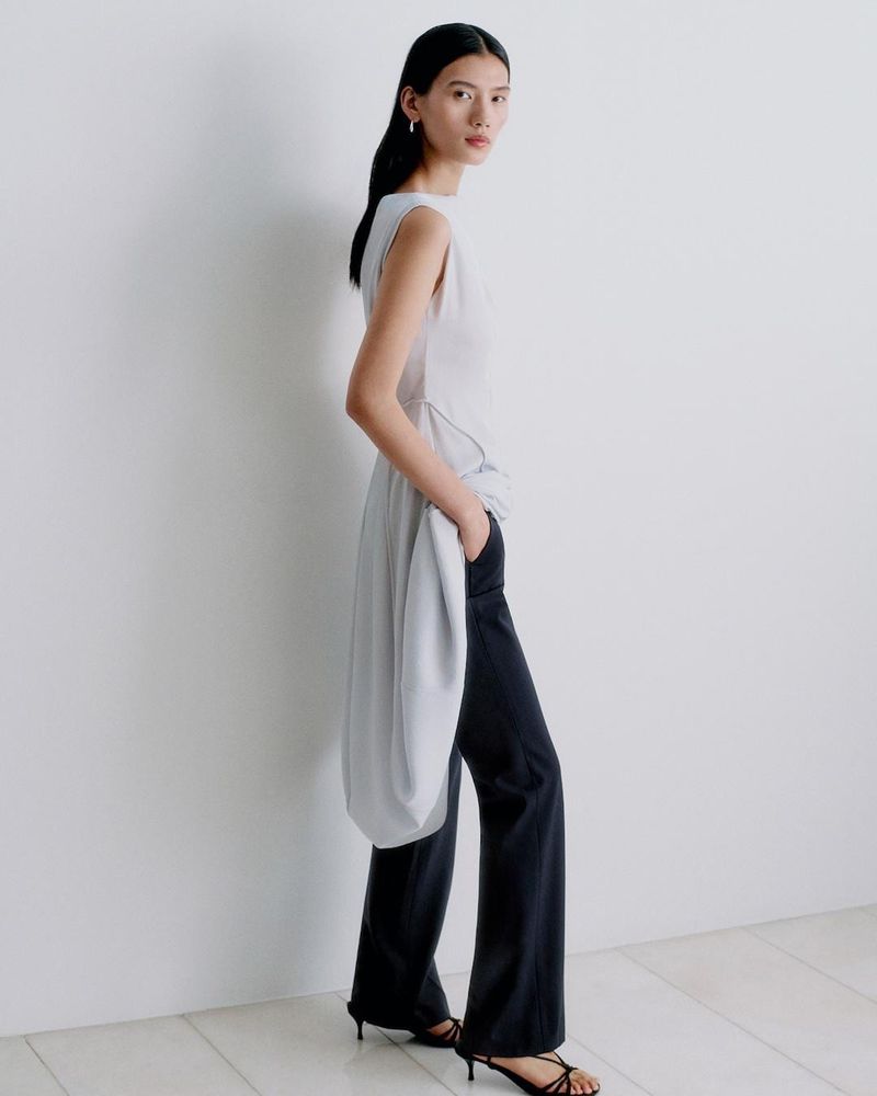 Model Jiayun Xie wearing a light grey draped sleeveless tunic and black wide-leg trousers against a white studio background.