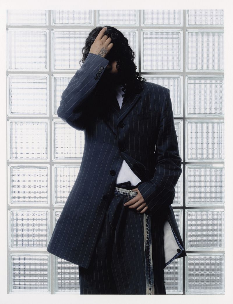 Person with long wavy hair poses in a pinstripe suit against a frosted glass block wall, with a white border for a modern editorial look.