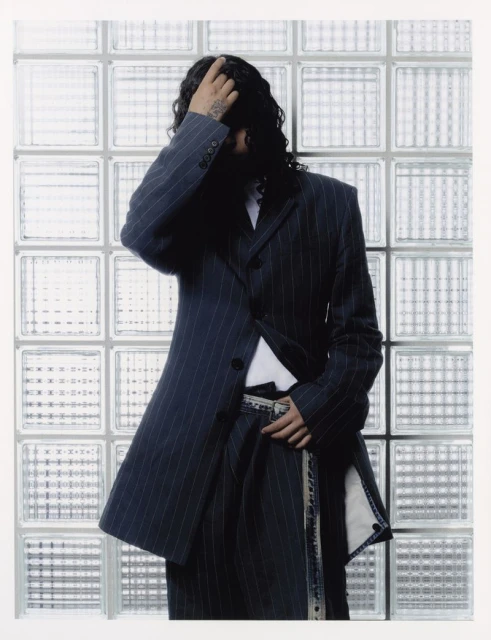Person with long wavy hair poses in a pinstripe suit against a frosted glass block wall, with a white border for a modern editorial look.