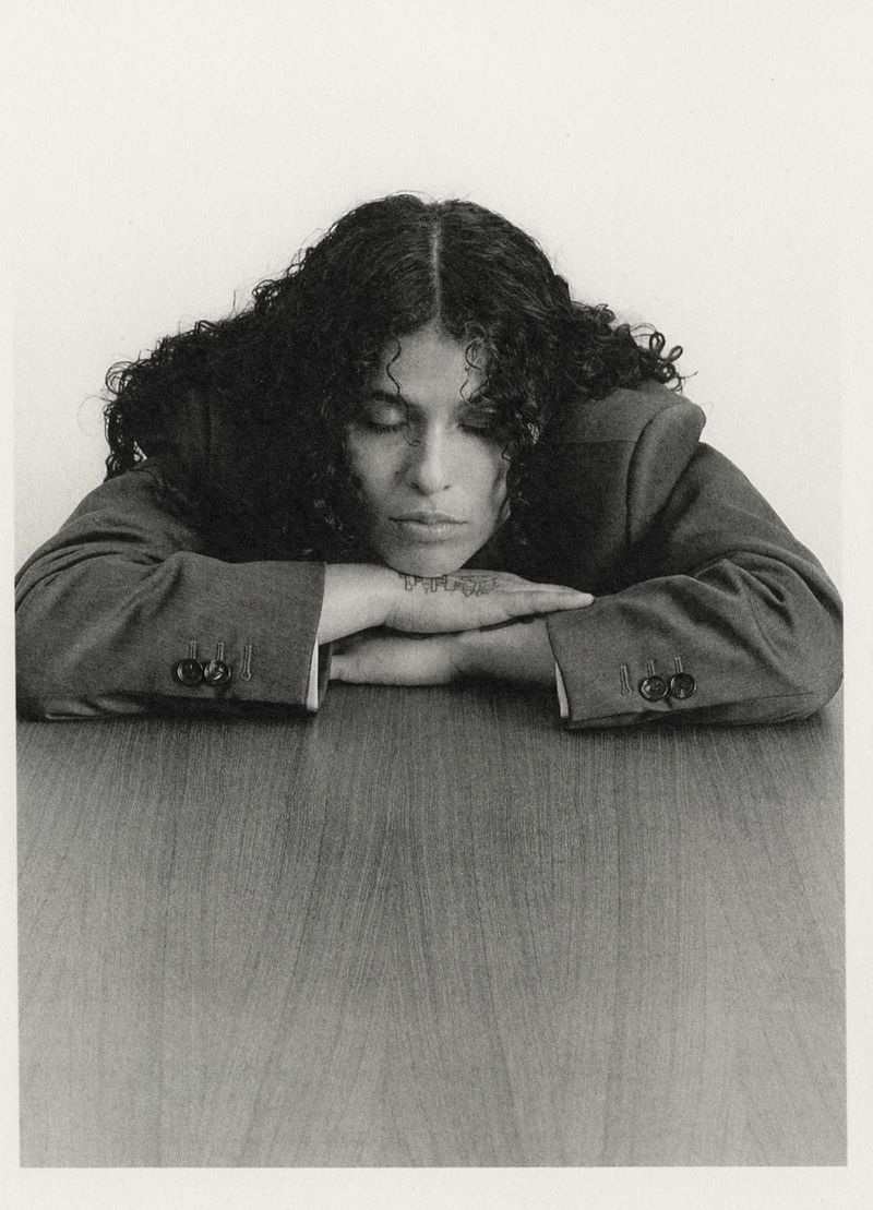 Woman with curly hair rests her chin on clasped hands on a table indoors against a white backdrop in a portrait-style studio shot.