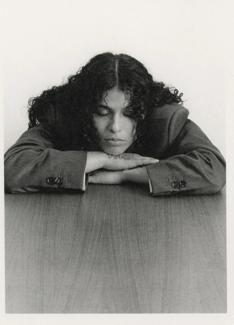 Woman with curly hair rests her chin on clasped hands on a table indoors against a white backdrop in a portrait-style studio shot.