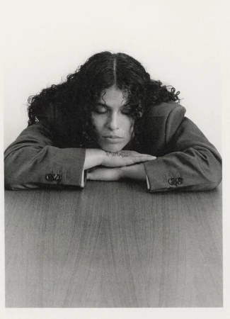 Woman with curly hair rests her chin on clasped hands on a table indoors against a white backdrop in a portrait-style studio shot.