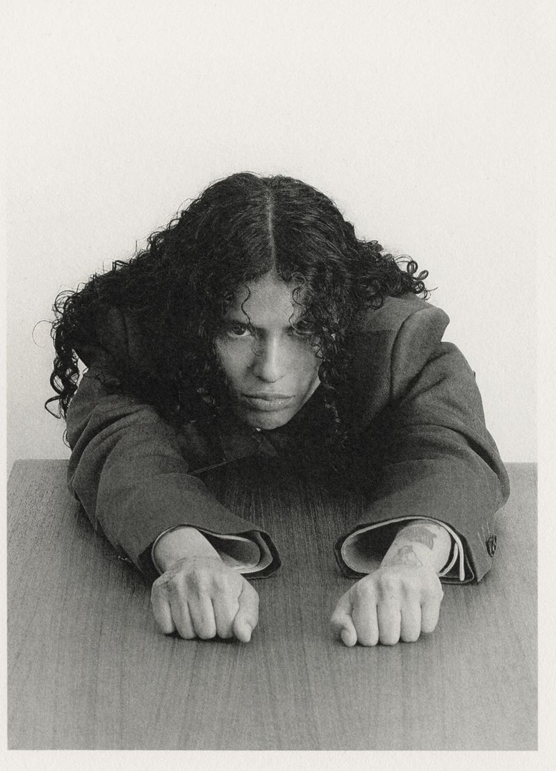 Monochrome portrait of a person with dark curly hair, wearing a suit, leaning on a wooden table, looking at the camera indoors.