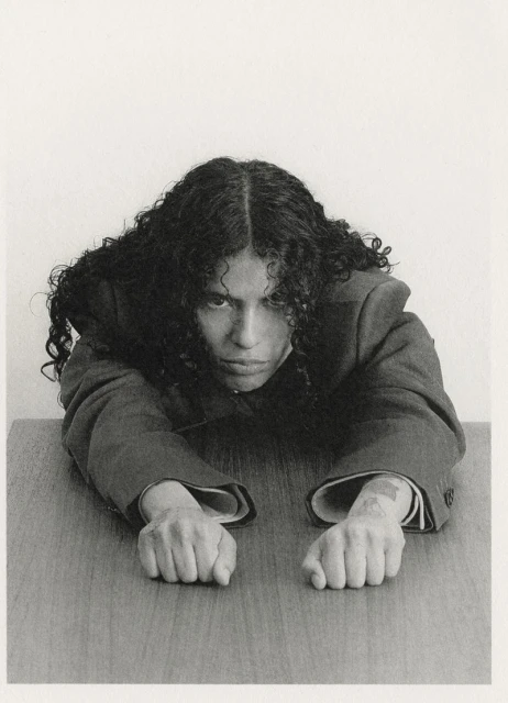 Monochrome portrait of a person with dark curly hair, wearing a suit, leaning on a wooden table, looking at the camera indoors.