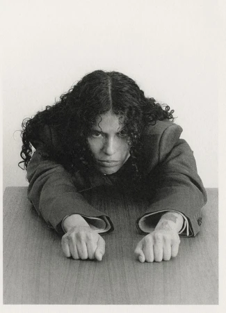 Monochrome portrait of a person with dark curly hair, wearing a suit, leaning on a wooden table, looking at the camera indoors.