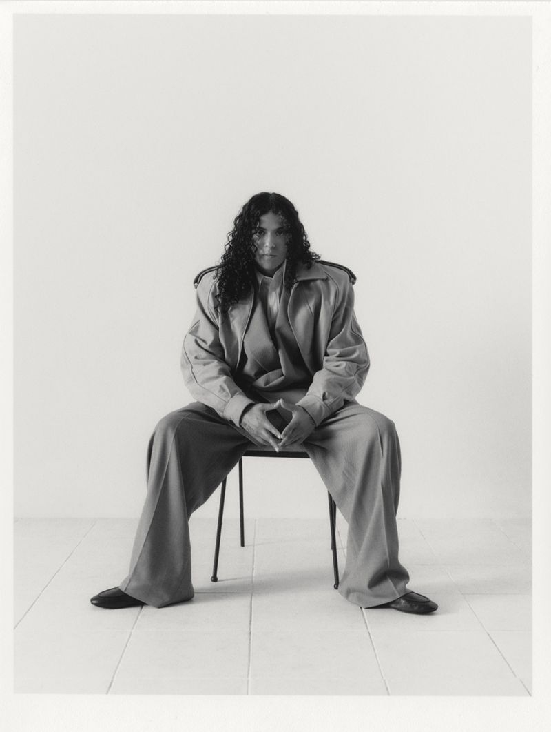 Black-and-white portrait of a person with long dark hair sitting on a chair wearing a blazer and wide-leg pants in a studio setting.