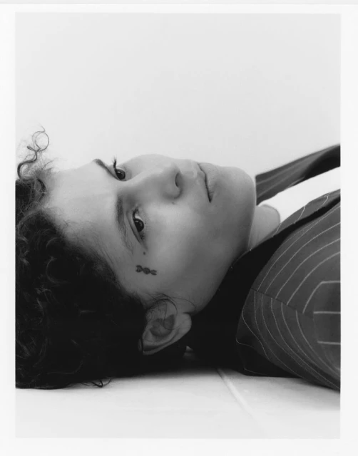 Monochrome portrait of a woman with short curly hair, lying down in pinstripe jacket, looking at the camera with a tattoo on her face.