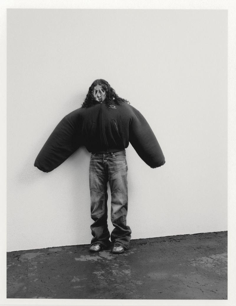 Person wearing an oversized black puffer jacket and jeans stands against a wall in a studio setting, creating an artistic and unusual image.