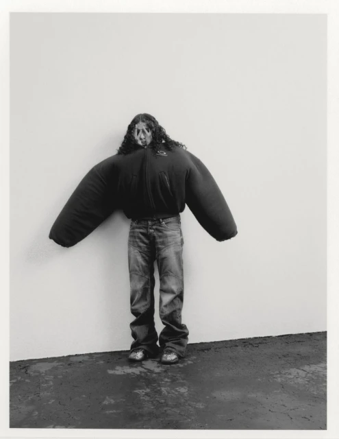 Person wearing an oversized black puffer jacket and jeans stands against a wall in a studio setting, creating an artistic and unusual image.