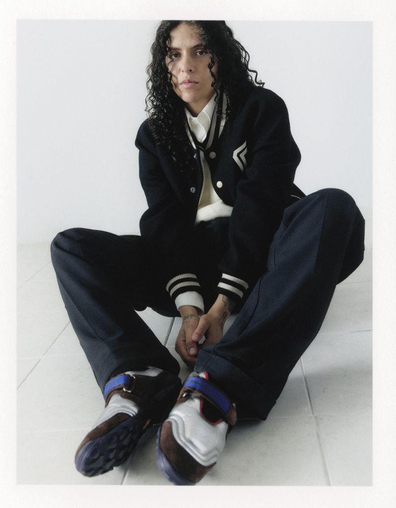 A young adult poses indoors wearing a black varsity jacket, cream-colored collared shirt, dark pants, and unique brown and white sneakers.