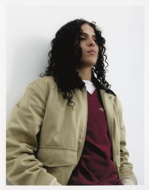Portrait of a young adult woman with curly dark hair wearing a beige jacket over a maroon v-neck shirt, standing in front of a white wall.