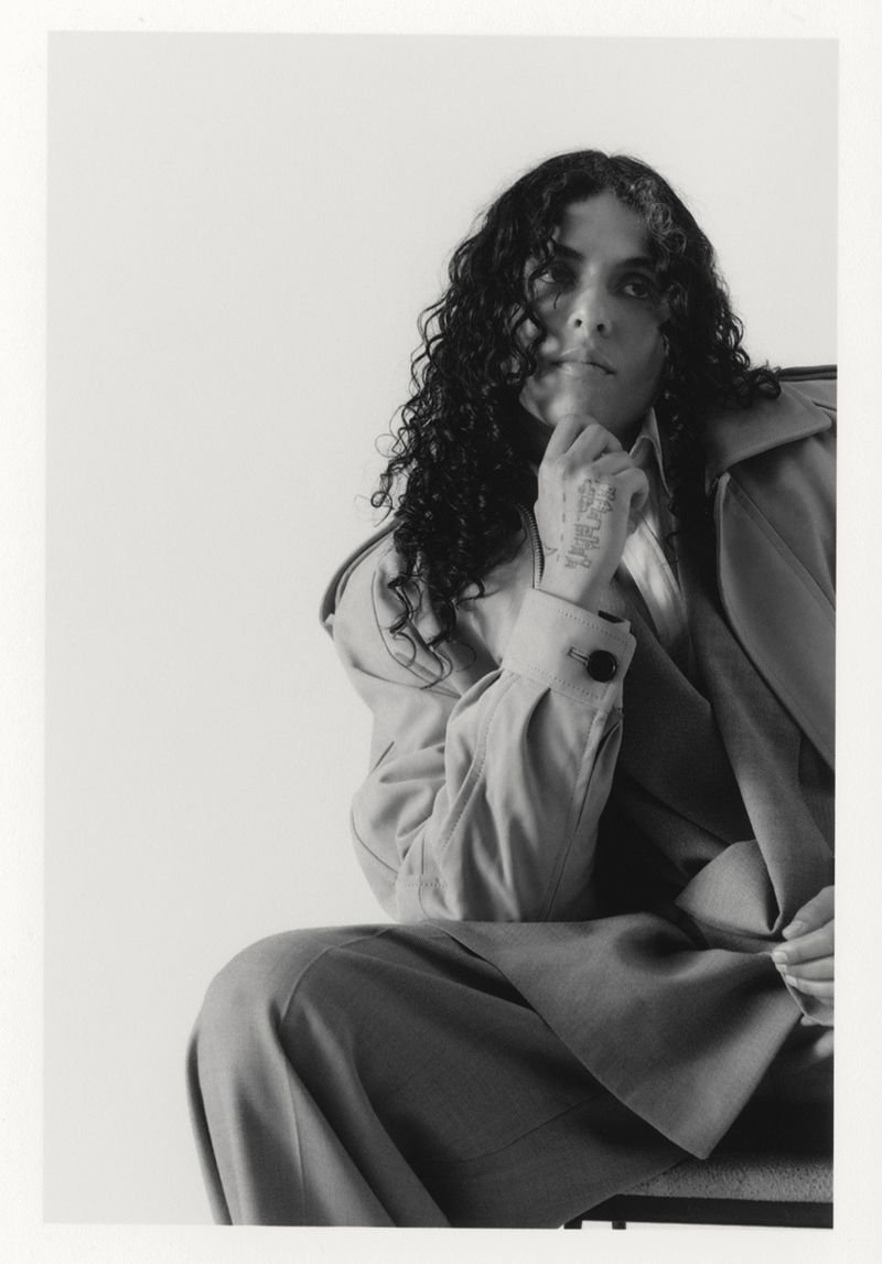 Monochrome portrait of a person with curly hair wearing a suit, seated indoors against a white backdrop with hand to chin.