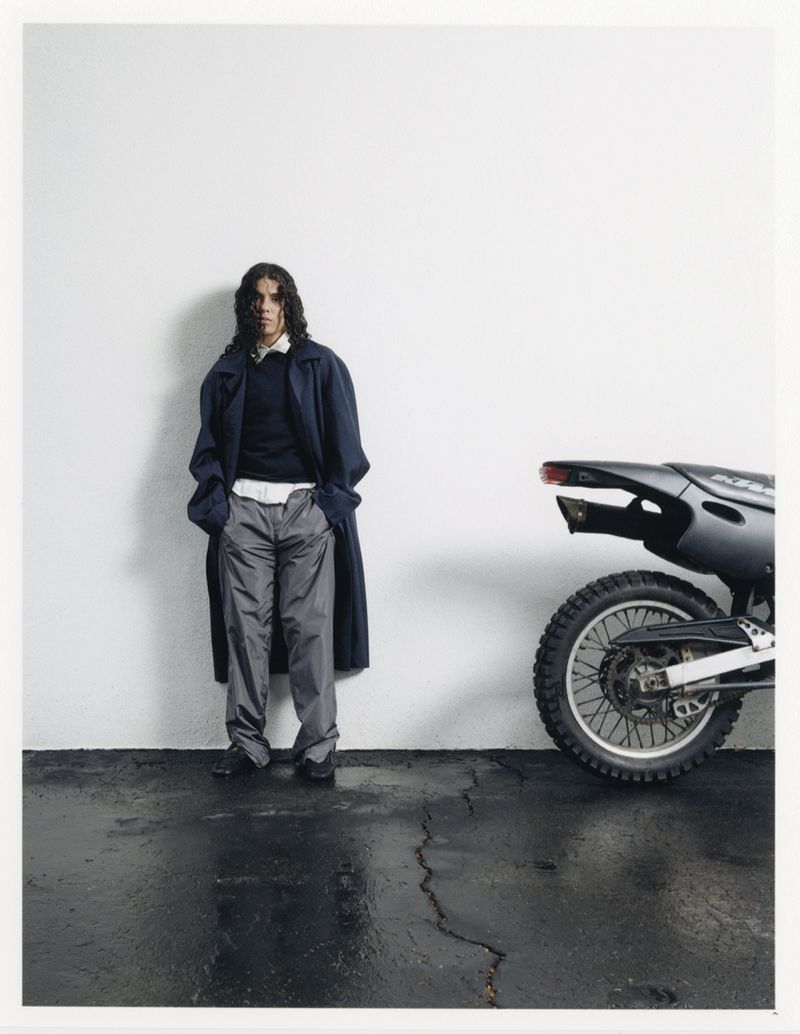 A person in a coat and grey pants stands next to a dark dirt bike on a wet black surface in front of a white wall.