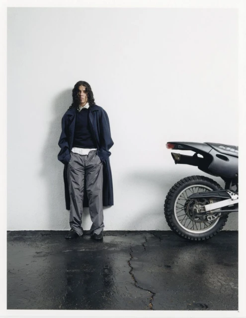 A person in a coat and grey pants stands next to a dark dirt bike on a wet black surface in front of a white wall.