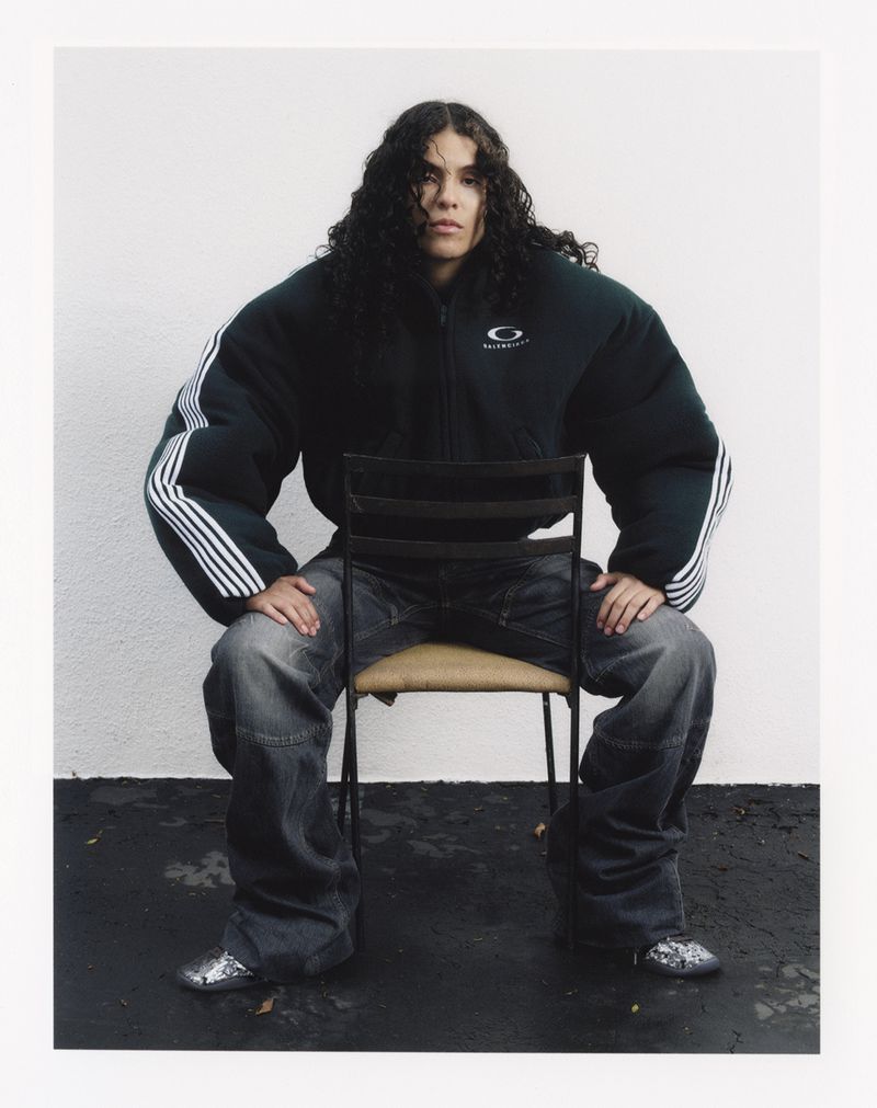 Person with long curly hair sits on a metal chair in an outdoor portrait, wearing an oversized green jacket and baggy jeans against a white wall.
