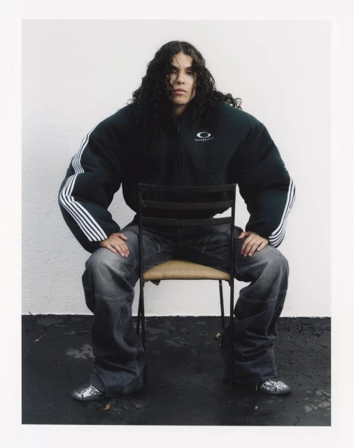 Person with long curly hair sits on a metal chair in an outdoor portrait, wearing an oversized green jacket and baggy jeans against a white wall.