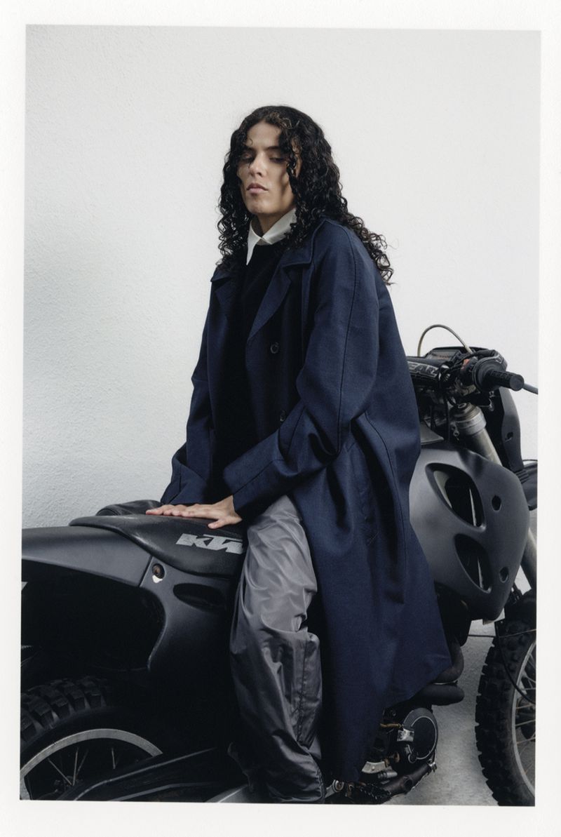 Adult with curly hair in long coat poses on a motorcycle in a studio with neutral backdrop and soft lighting, fashion editorial style.