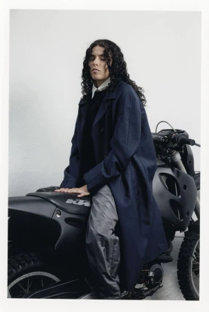 Adult with curly hair in long coat poses on a motorcycle in a studio with neutral backdrop and soft lighting, fashion editorial style.