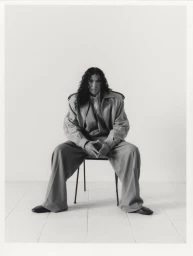 070 Shake seated in an oversized tailored suit and wide-leg trousers on a minimalist chair against a white studio backdrop.
