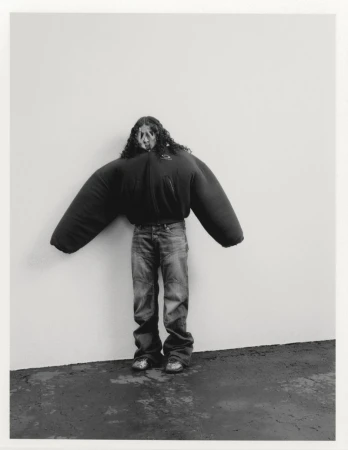 070 Shake posing in an exaggerated oversized sculptural black jacket and jeans against a minimal white studio wall.