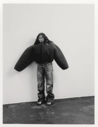 070 Shake posing in an exaggerated oversized sculptural black jacket and jeans against a minimal white studio wall.