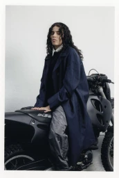 070 Shake wearing a long navy blue coat and grey trousers seated on a black motorcycle against a plain white wall.