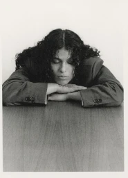 Portrait of 070 Shake resting head on crossed arms at a wooden table wearing a dark tailored blazer.