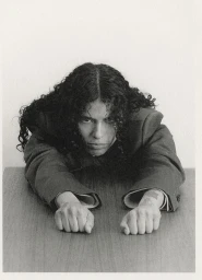 A person with long dark curly hair leaning over a wooden table, wearing a dark blazer.