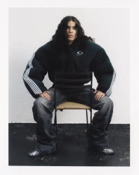 Musician 070 Shake seated backwards on a chair wearing an exaggerated oversized dark puffer jacket and baggy washed grey jeans.