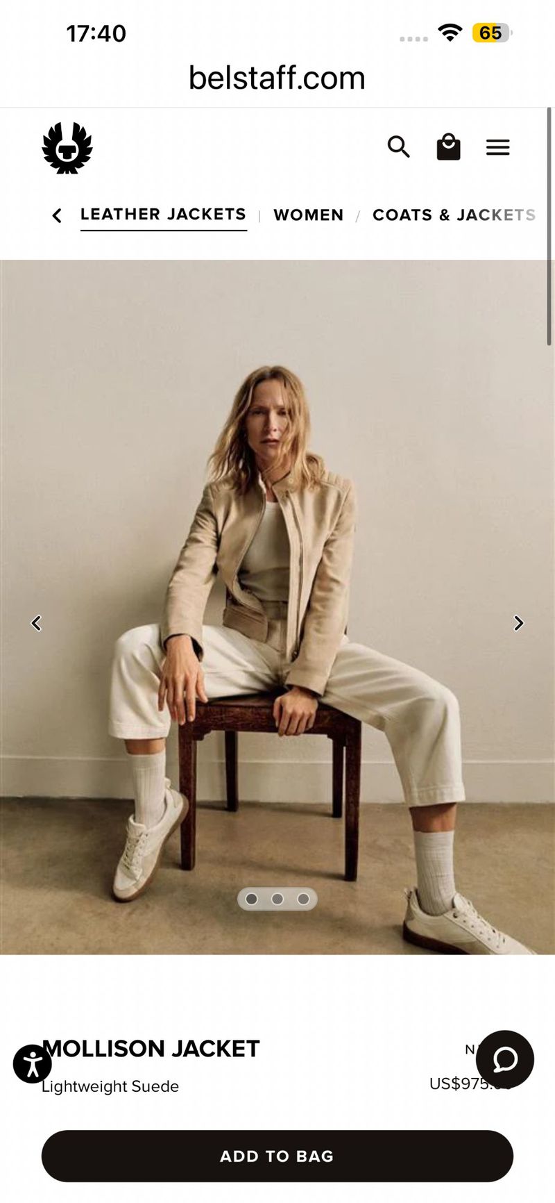 Model seated on a wooden chair wearing a beige jacket and light trousers against a neutral background