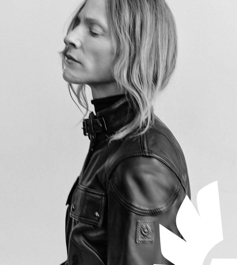 Profile portrait of a model wearing a black Belstaff leather motorcycle jacket with a buckled collar against a light studio backdrop.