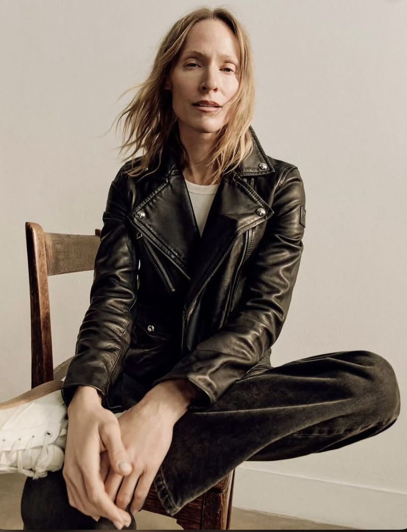 Lindsay Sullivan sitting on a wooden chair wearing a black leather biker jacket, dark trousers, and white sneakers.