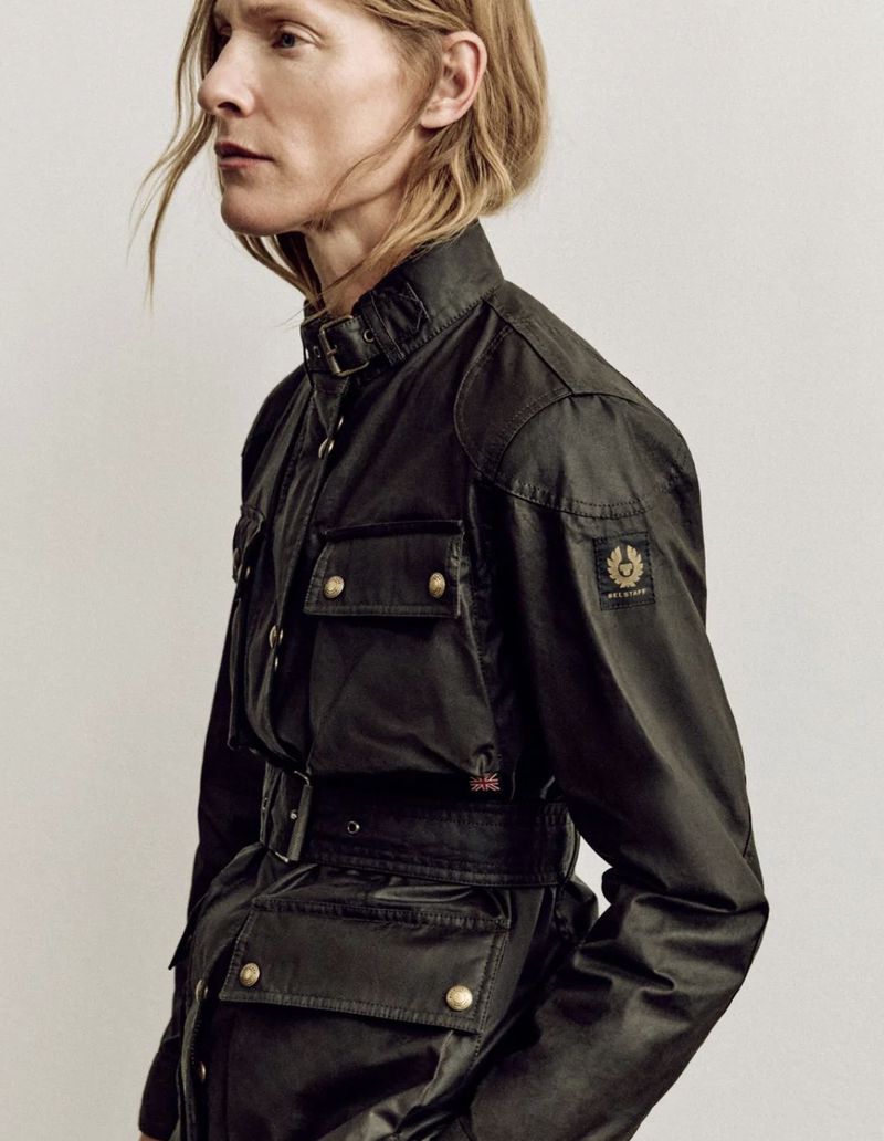 Lindsay Sullivan wearing a belted black waxed jacket with brass snap buttons and a high collar against a plain studio background.