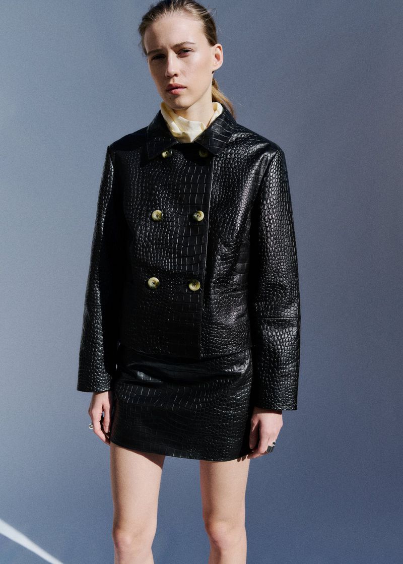 Model Liva Been wearing a black crocodile-embossed jacket and matching mini skirt against a cool grey studio backdrop.