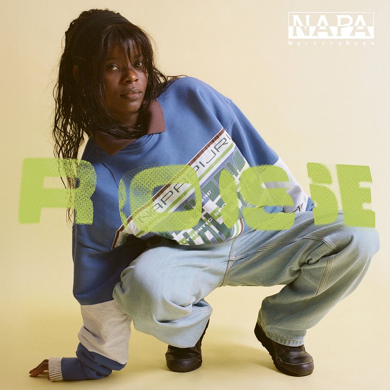 Black woman crouching against a pale backdrop wearing a blue sweatshirt and light wash jeans