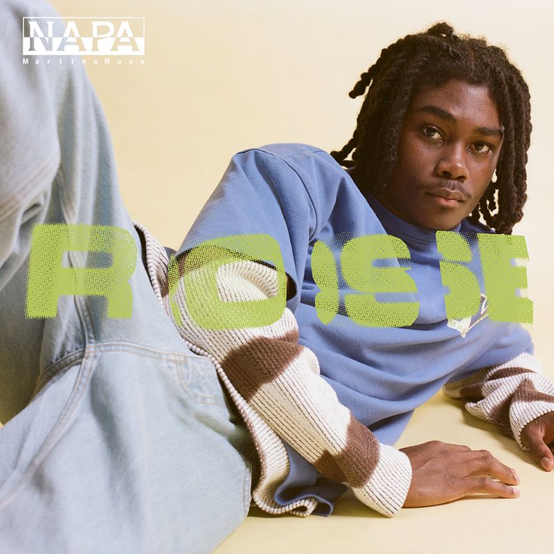 A young Black man with dreadlocks reclines in light blue jeans, a blue shirt, and a striped knit sleeve on a pale surface.