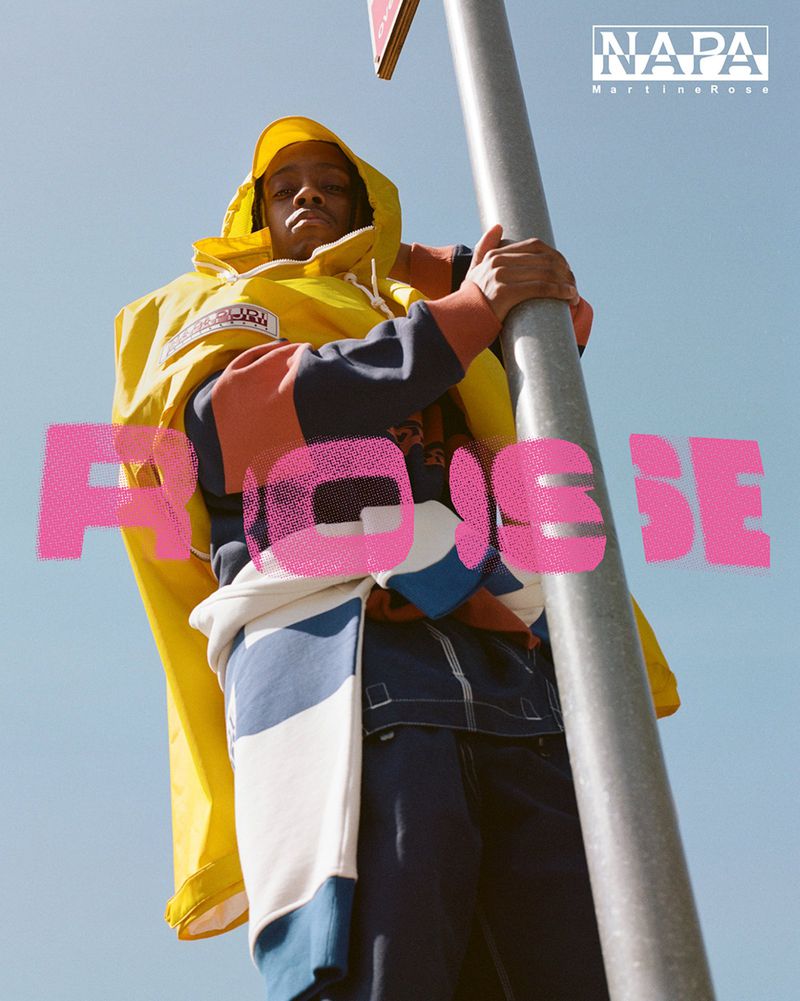 Low angle of a man leaning on a pole wearing a bright yellow Martine Rose x Napapijri hooded jacket under a clear sky.