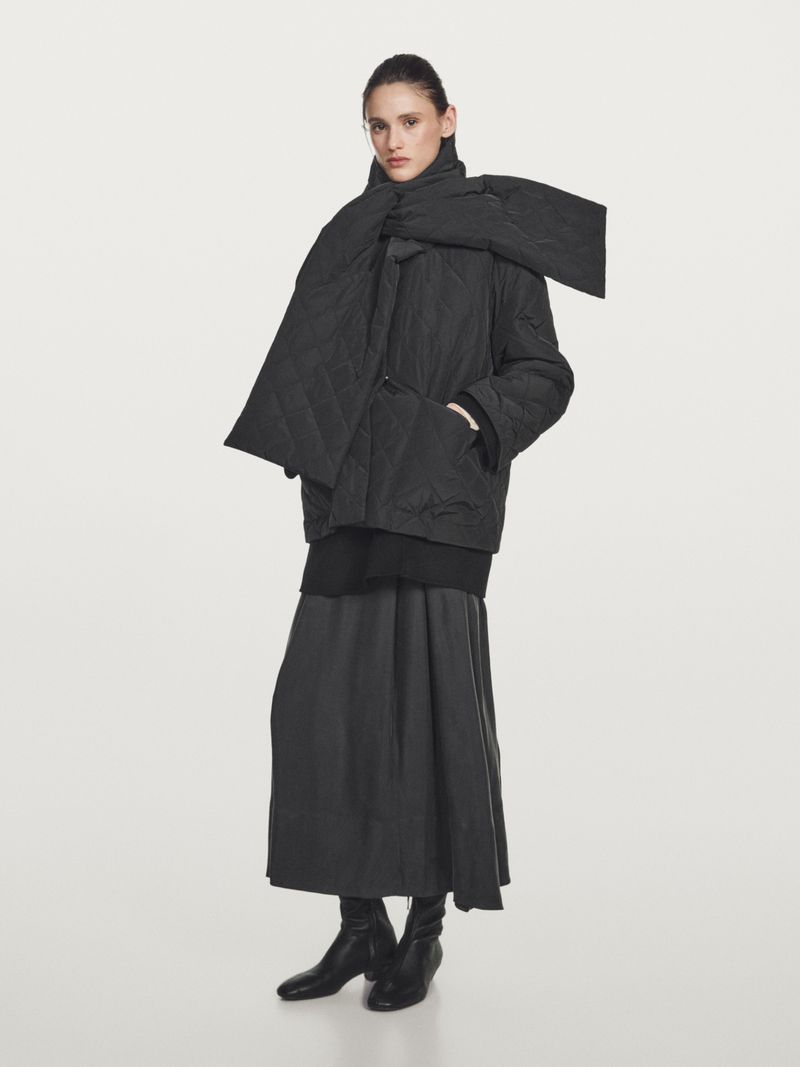 Model Aleyna Fitzgerald wears an oversized black quilted jacket, matching scarf, long skirt, and black boots against a studio backdrop.