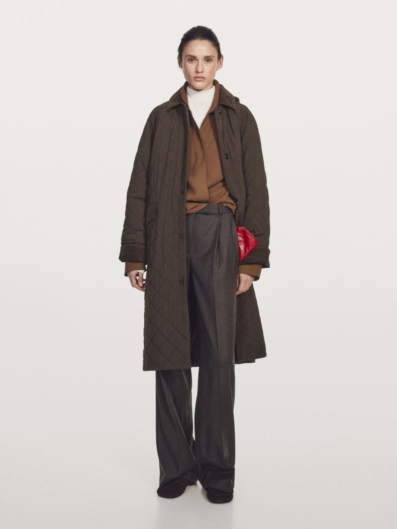 Model Aleyna Fitzgerald wearing a long brown quilted coat and dark pleated wide-leg trousers against a neutral studio backdrop.