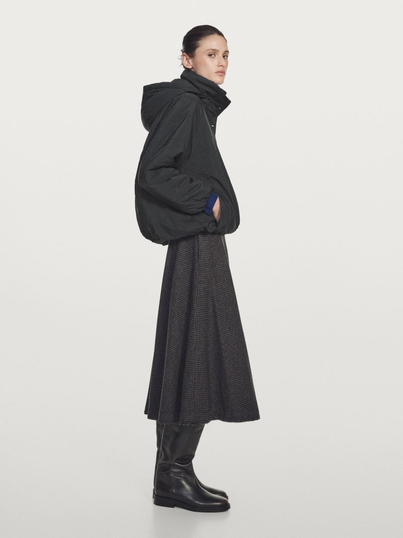 Aleyna Fitzgerald wearing a black hooded jacket, dark flared midi skirt, and black leather boots against a light grey seamless backdrop.