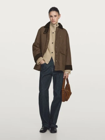 Model in brown jacket and blue jeans holding a brown suede handbag against a neutral white seamless background.